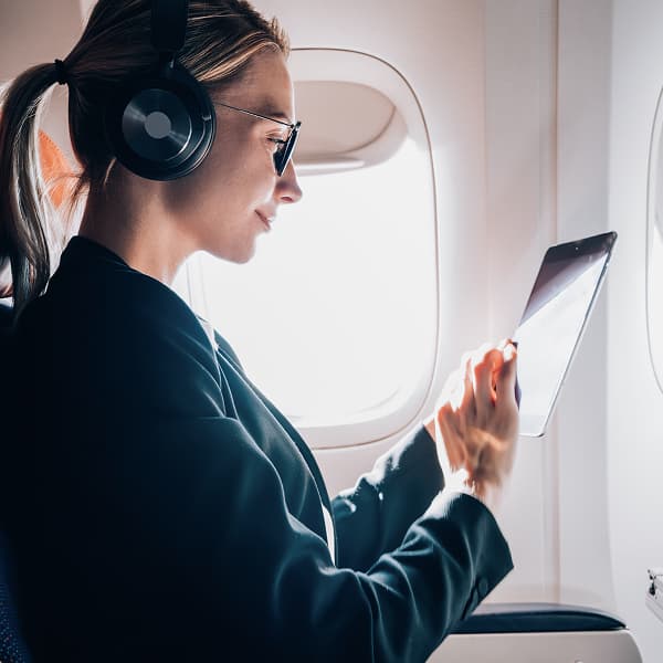 A woman in a black jacket and headphones uses a tablet on an airplane. Sunlight streams through the window, creating a calm, focused atmosphere.