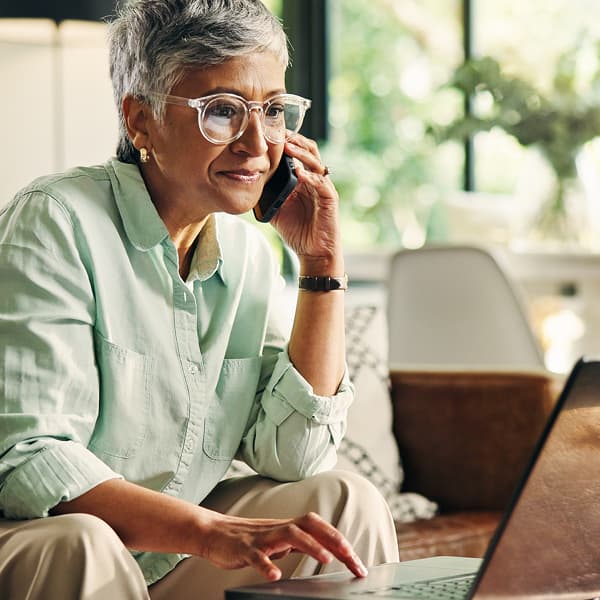 Elderly woman with short gray hair and glasses multitasking at home, talking on a phone while typing on a laptop. She appears focused and content.