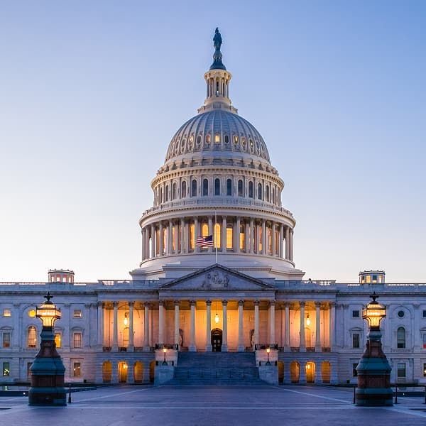 The U.S. Capitol building at dusk, illuminated with warm lights. The dome stands prominent against the clear sky, conveying a sense of grandeur and history.