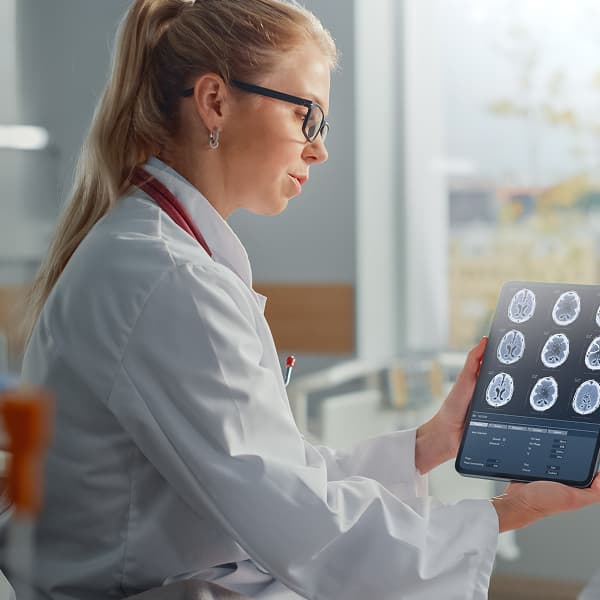 A female doctor in a white coat examines brain scans on a tablet with a focused expression. She sits beside a patient, creating a calm, professional atmosphere.
