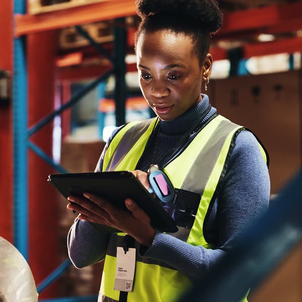A focused worker in a high-visibility vest uses a tablet in a warehouse setting. Shelves line the background, conveying an organized, industrious atmosphere.