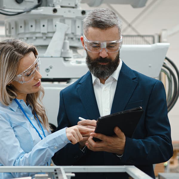Two professionals in safety goggles discuss a tablet in an industrial setting, standing by machinery, conveying focus and collaboration.