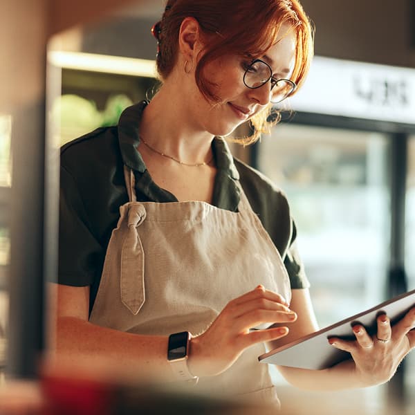 A woman with red hair, wearing glasses and a beige apron, is holding a tablet in a bright café. She looks focused and content.