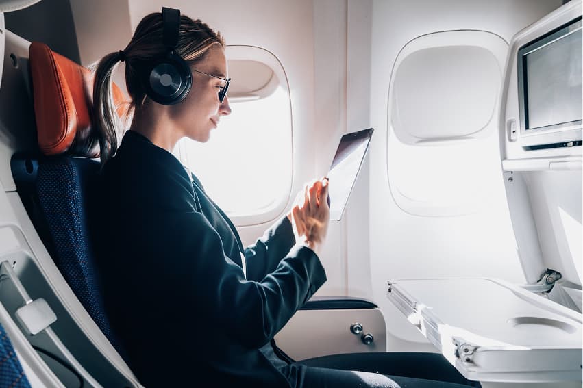 A woman sitting in an airplane seat, looking at a tablet with a thoughtful expression. The interior of the plane is visible around her, including the seat and overhead compartments.