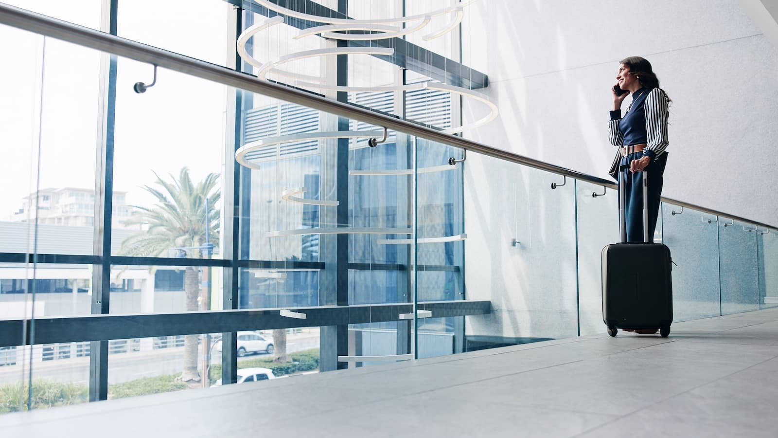 A woman in a striped blazer stands with a suitcase in a sunlit airport terminal, talking on her phone. Modern design, glass windows, and palm trees outside.