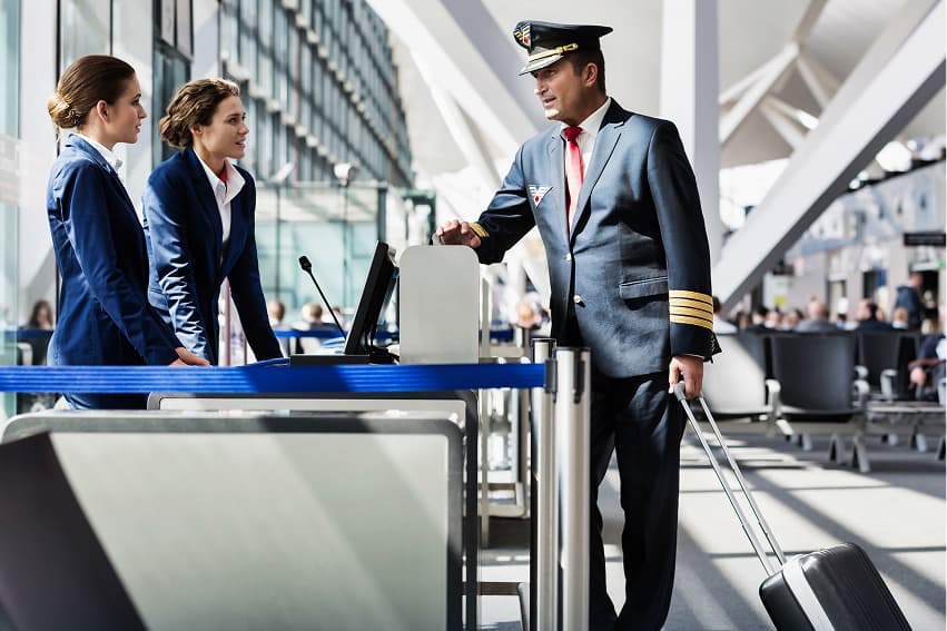 Airline pilot with a suitcase talking to two flight attendants at an airport gate. They appear focused and professional, set in a bright terminal.