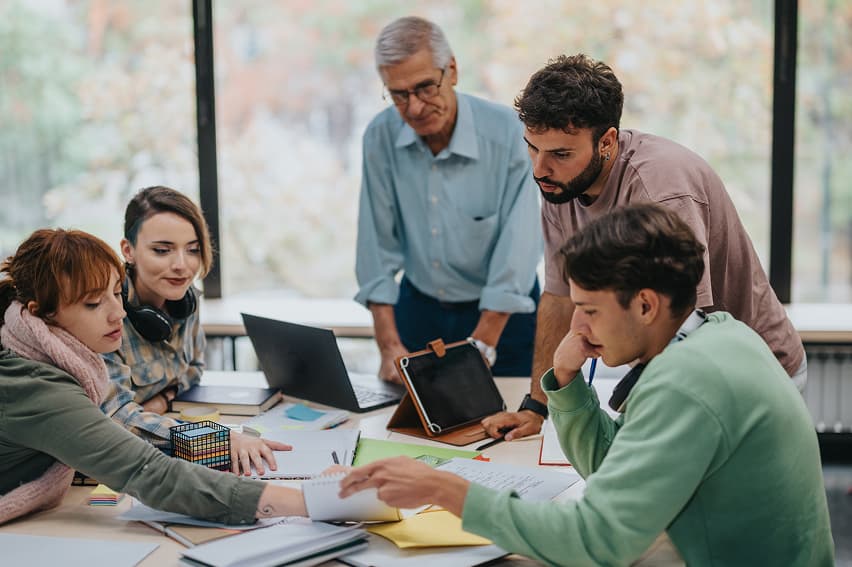 A diverse group of five people collaborate at a table with laptops and papers, conveying teamwork and focus in a bright, modern office setting.