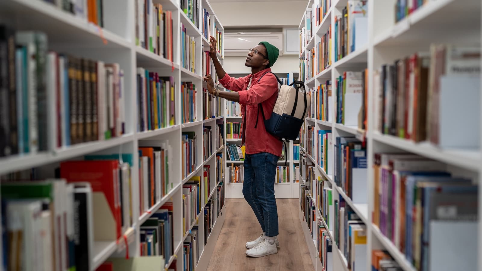 A person wearing a green beanie and red shirt browses books on high shelves in a library. They carry a backpack, conveying focus and curiosity.