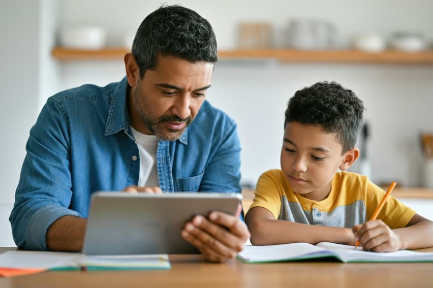 Man and child doing homework at a table. The man, in a denim shirt, uses a tablet, while the child, in a yellow shirt, writes in a notebook.
