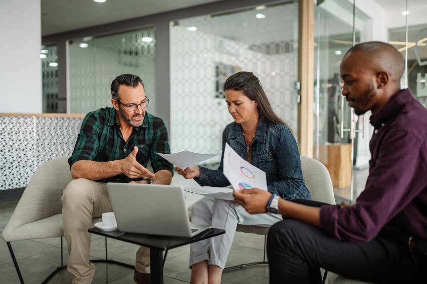 Three people sit in an office, engaged in a discussion. They focus on documents and a laptop, conveying a collaborative and professional atmosphere.