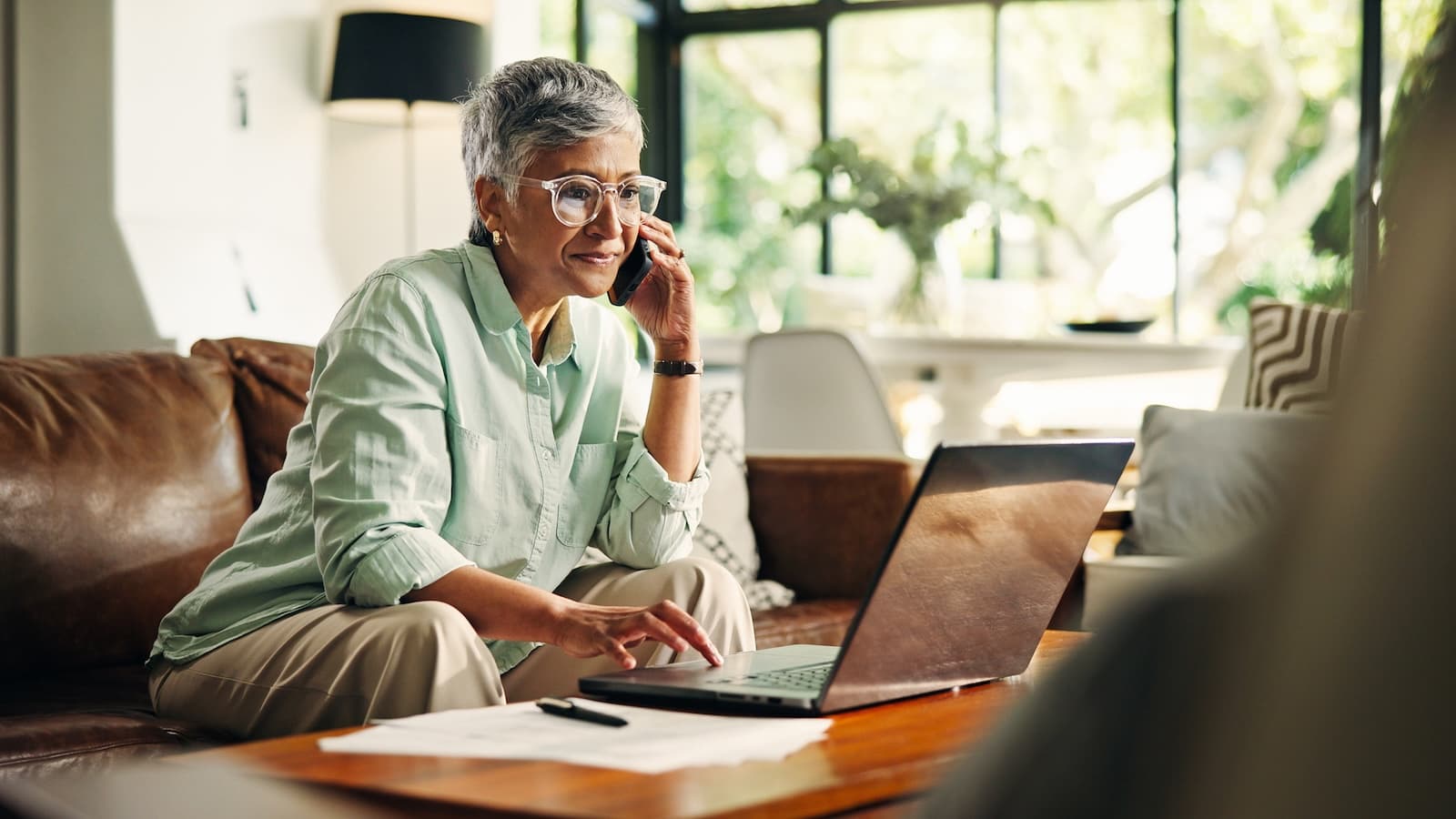 Elderly woman with short gray hair, smiling while talking on phone and using a laptop in a bright living room. Sunlight streams through large windows behind her.