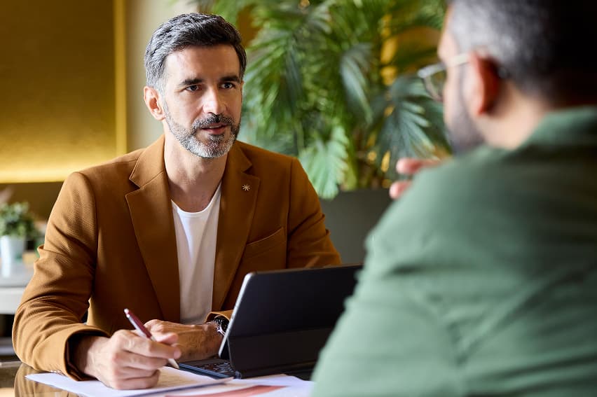 A man in a brown blazer speaks attentively to another person at a table with a laptop. Background shows lush green plants, creating a calm atmosphere.