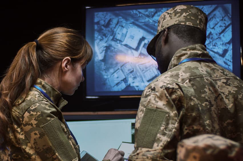 Two military personnel in camouflage uniforms analyze a digital map on a screen in a dimly lit room, conveying focus and strategy.