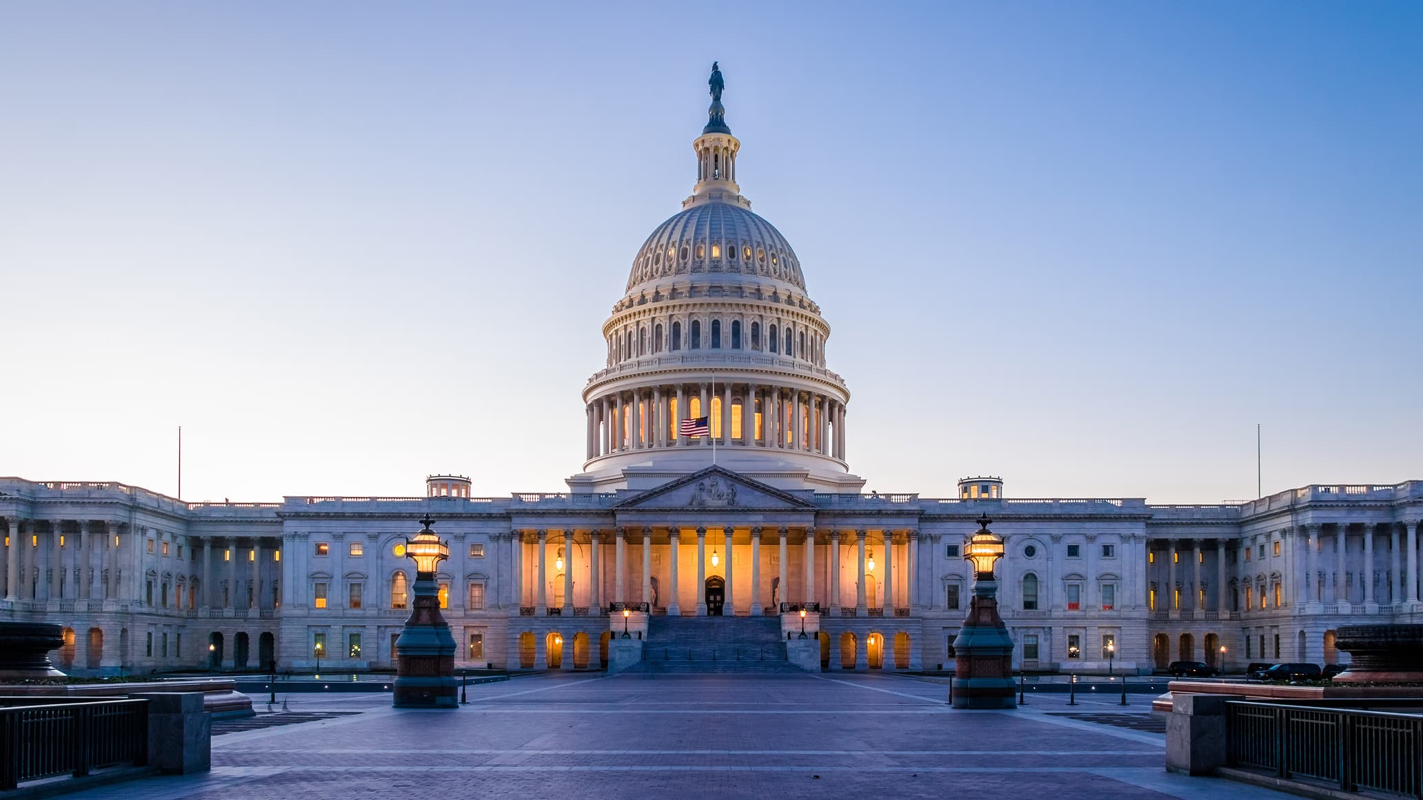 The U.S. Capitol building at dawn, with a clear sky backdrop. Its dome is illuminated by warm lights, conveying a sense of grandeur and tranquility.