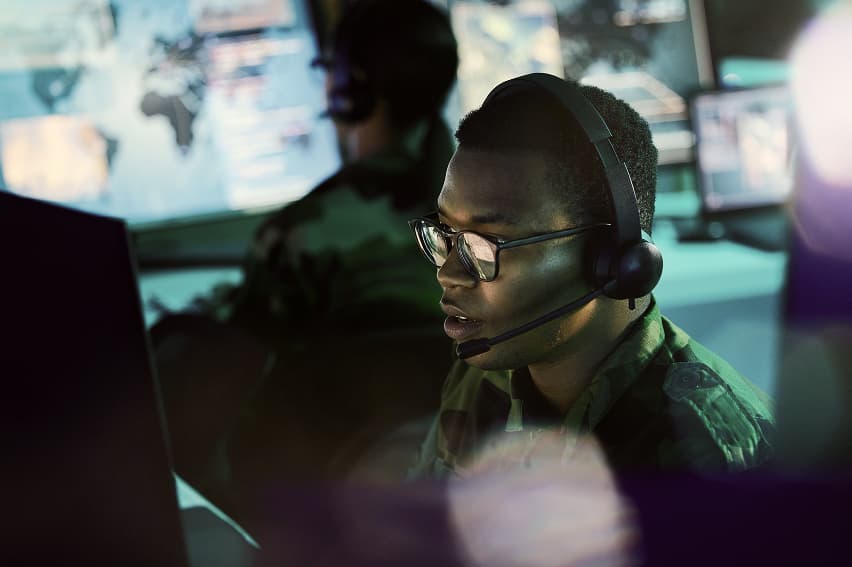 A person wearing glasses and a headset focuses intently on a computer screen in a dimly lit room with multiple monitors displaying maps and data, suggesting a high-tech or military operation.
