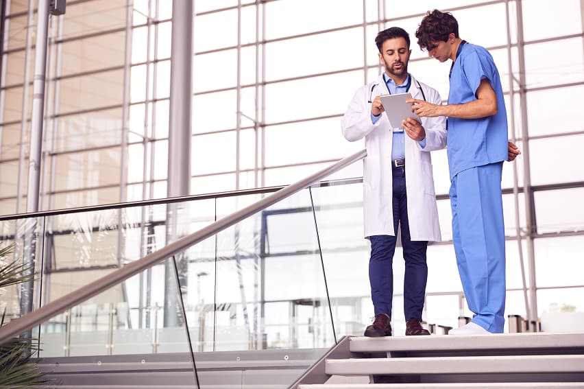 Two healthcare professionals, one in a white coat and the other in blue scrubs, discuss a tablet in a modern hospital with large windows.