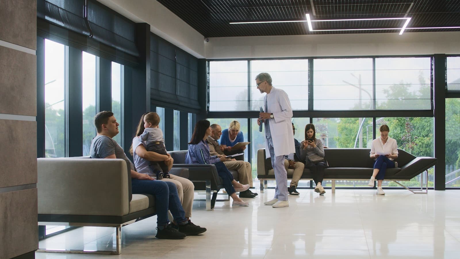 A doctor in a white coat speaks with seated patients in a bright, modern waiting room. Some read, while others look on attentively, creating a calm atmosphere.