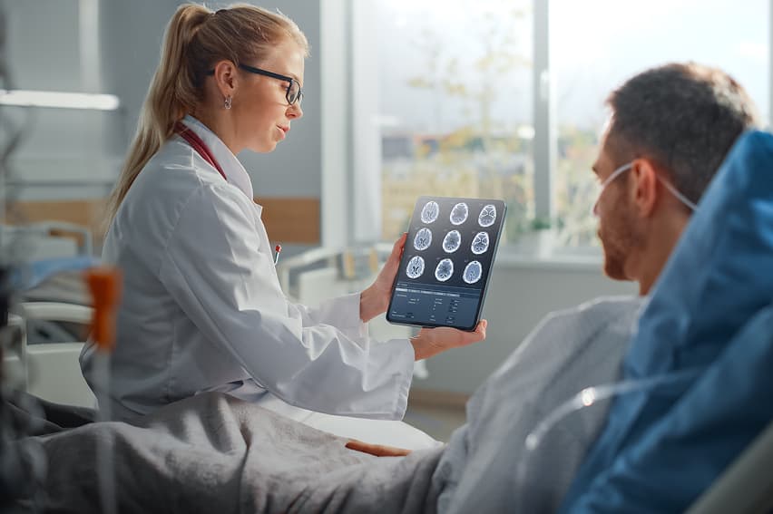 A doctor in a white coat and stethoscope shows a male patient brain scan images on a tablet. The setting is a well-lit hospital room, conveying a professional and informative tone.