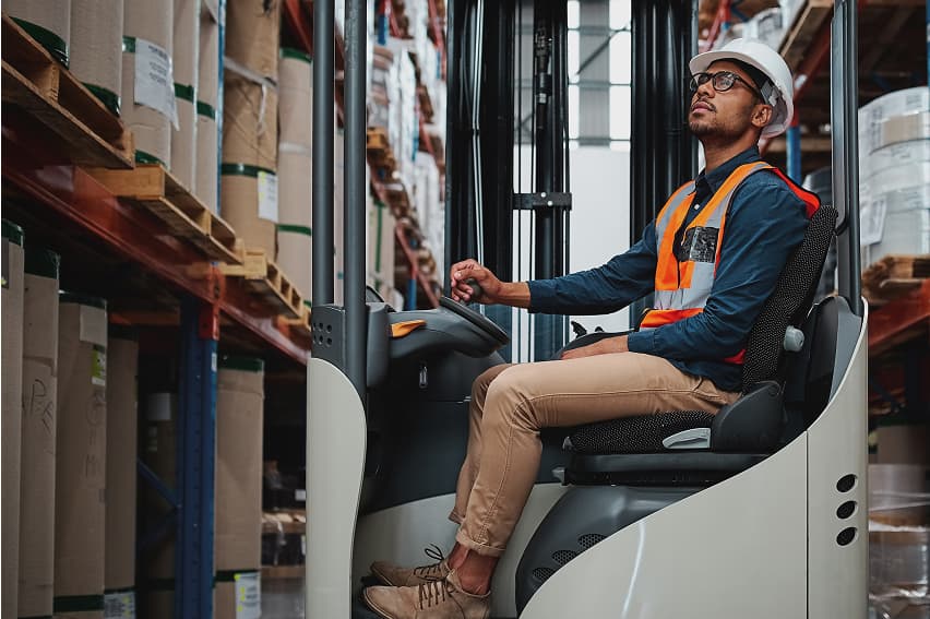 Worker in a warehouse operates a forklift, wearing a white hard hat and orange safety vest. Shelves filled with boxes line the background.