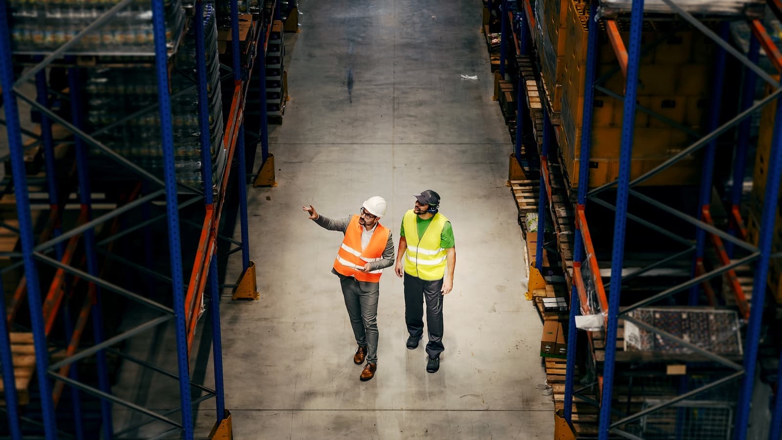 Two people in high-visibility vests walk down an aisle in a warehouse. One gestures while they discuss logistics, surrounded by shelving and pallets.