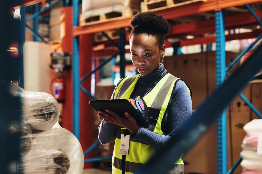 Woman in a high-visibility vest uses a tablet in a warehouse, surrounded by shelves with boxes and large rolls. The setting appears organized and focused.
