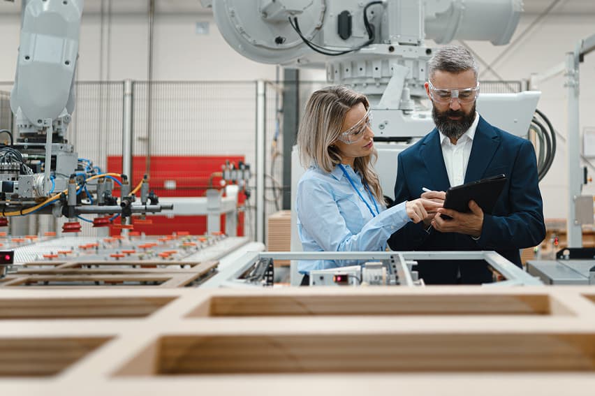 Two people in safety glasses examine a tablet in an industrial setting with robotic arms. They appear focused, discussing machinery operations.