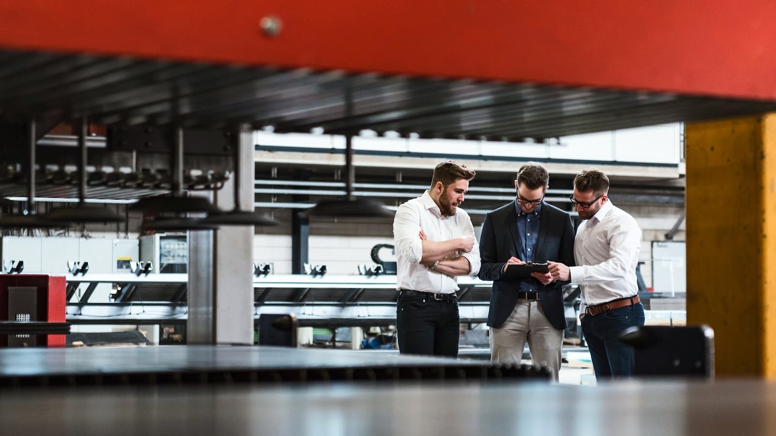Three men stand in a factory, examining a tablet. They're dressed in business casual attire, conveying a tone of focus and collaboration. Machinery surrounds them.