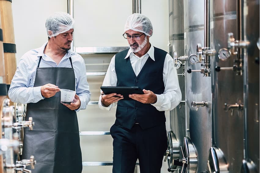 Two men wearing hairnets inspect equipment in a brewery or industrial kitchen. One holds a clipboard, the other holds a notepad, engaged in discussion.