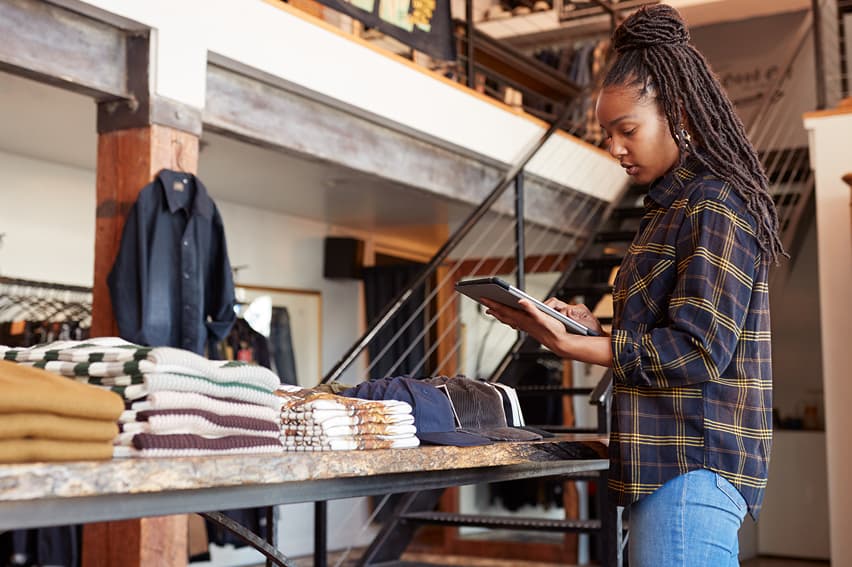 A woman in a plaid shirt uses a tablet in a clothing store. Folded sweaters and shirts are on a table. The setting feels modern and orderly.