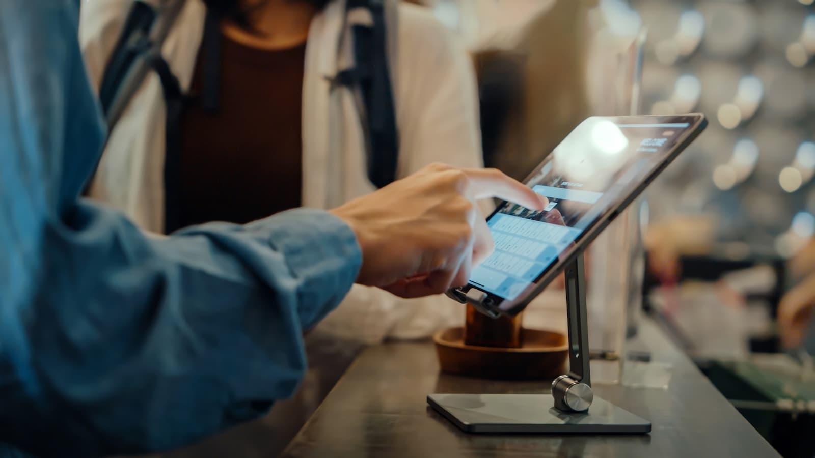Close-up of a person's hand using a touchscreen tablet on a stand at a store counter. Another person stands nearby. The setting is modern and busy.