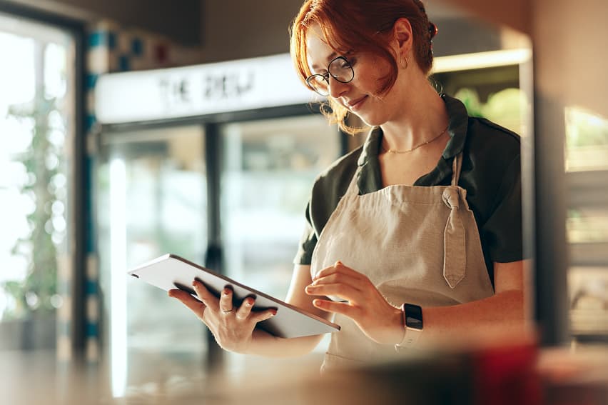 A woman with red hair and glasses, wearing an apron, stands in a cafe using a tablet. The setting is bright and casual, conveying a focused atmosphere.